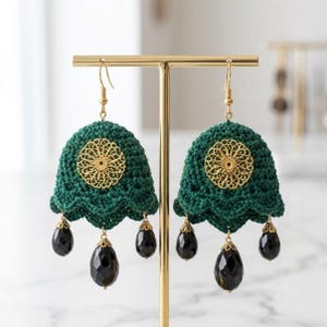 May include: A pair of dark green crochet earrings featuring a gold-toned filigree circular accent at the center and three dangling black teardrop-shaped beads suspended from the scalloped bottom edge, hanging from gold-toned metal hooks.