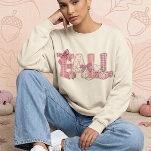 May include: A cream-coloured crewneck sweatshirt featuring the word FALL in large, pink, plaid-patterned letters. The design is decorated with pink bows, small floral clusters, a heart shape, and a small pumpkin graphic on the right side.