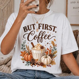 Könnte beinhalten: Ein weißes Kurzarm-T-Shirt mit einem Grafikdruck und dem Text "BUT FIRST, Coffee" in brauner Schrift. Das Design zeigt eine Kaffeetasse mit Schlagsahne, umgeben von Kürbissen, herbstlichen Blättern und Tannenzapfen.
