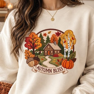 May include: A cream-colored crewneck sweatshirt featuring an embroidered circular design of a cabin in a forest with autumn trees, acorns, and a pumpkin. Below the scene, a banner displays the text AUTUMN BLISS in brown capital letters.