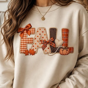 May include: A cream-colored crewneck sweatshirt featuring the word FALL appliqued in various textured fabrics. The letters include orange gingham, maple leaf patterns, brown corduroy, and plaid, accented with satin bows, a heart, and a pumpkin.