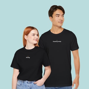 May include: Two black short-sleeved t-shirts. The smaller shirt features the word Arising in white text with a small gold icon above it. The larger shirt features the words Passing away in white text with a small gold icon above it.