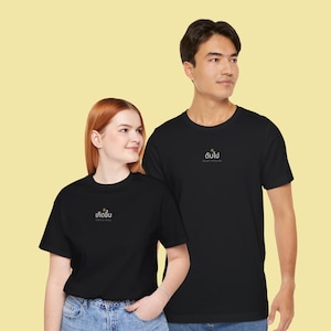 May include: Two black short-sleeved crew neck t-shirts. The shirt on the left features the Thai text "เกิดขึ้น" with a small gold icon above it. The shirt on the right features the Thai text "ดับไป" with a small gold icon above it.