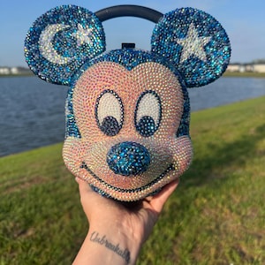 Puede incluir: Bolso con forma de Mickey Mouse adornado con pedrería iridiscente. El bolso tiene un asa negra y orejas azules, una con una luna creciente, la otra con una estrella. La cara es rosa con ojos negros y una nariz azul.