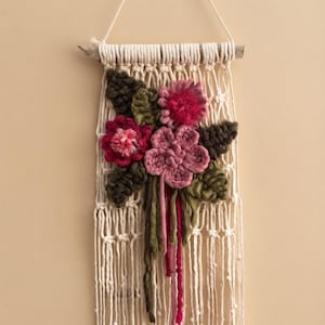 Op de afbeelding: Een macramé wandhanger van off-white katoenkoord geknoopt aan een houten tak. Het werk is versierd met driedimensionale garenbloemen in roze en magentatinten, vergezeld door donker olijfgroene garenbladeren en loshangende draden aan de onderzijde.