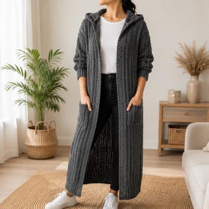 May include: A long, dark gray hooded cardigan featuring a vertical ribbed knit texture, long sleeves, and two large front patch pockets. The open-front garment is worn over a plain white t-shirt and black pants, paired with white sneakers.