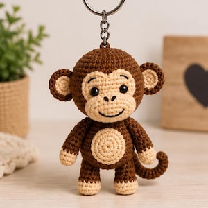 May include: A brown and beige crocheted monkey keychain featuring a round head with small ears, a light-colored face, a circular belly patch, and a curled tail. The monkey is attached to a silver-toned metal split ring by a short chain.