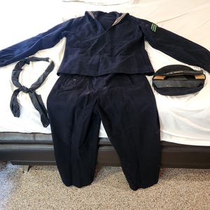 Puede incluir: Un conjunto de uniforme de marinero azul marino oscuro que consta de una camisa de manga larga con cuello tradicional y tres rayas verdes en la manga izquierda, pantalones a juego, un pañuelo negro y una gorra negra con el texto U.S. NAVY.