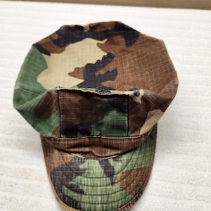 May include: A military-style patrol cap featuring a woodland camouflage pattern in shades of brown, green, and black. The cap is constructed from a grid-textured fabric and includes a flat, rounded top with a structured brim extending from the front.