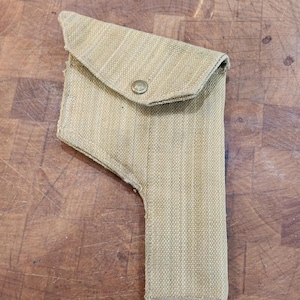 May include: A tan canvas holster featuring a structured shape with a fold-over flap secured by a single metal snap button. The holster is constructed from a durable, woven fabric material and has a reinforced bottom edge with visible stitching.