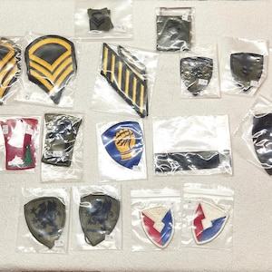 May include: A collection of various military-style embroidered patches in individual clear plastic bags. The assortment includes chevron rank insignia, shield-shaped unit patches with diverse emblems, and rectangular bars in colors like olive, black, blue, and gold.