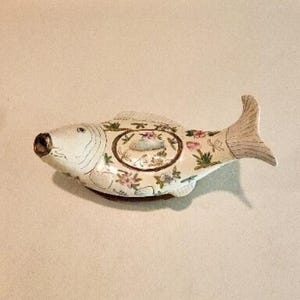Puede incluir: Un recipiente de cerámica con tapa en forma de pez de color blanco hueso. El exterior presenta motivos florales pintados a mano en tonos rosa, verde y marrón. Una pequeña figura de pez en relieve se encuentra centrada sobre la tapa.