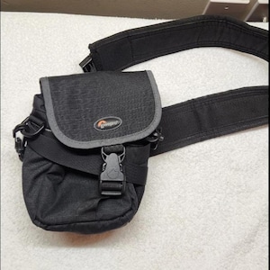 May include: A black Lowepro camera bag featuring a textured fabric exterior, a fold-over flap with a gray trim, and a central plastic buckle closure. The bag includes an adjustable black shoulder strap with a padded section for carrying comfort.