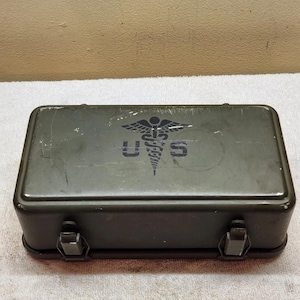Puede incluir: Una caja rectangular de suministros médicos de metal verde oliva con dos cierres frontales. La tapa presenta un símbolo de caduceo negro impreso sobre las letras U.S. La superficie muestra rasguños menores y signos de desgaste.