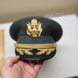 May include: A dark green military service cap featuring a gold-colored United States Army emblem on the front. The cap includes a textured black band, a gold chin strap, and gold-embroidered oak leaf detailing along the brim of the visor.