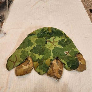 Puede incluir: Una funda de camuflaje para casco con un patrón de hojas en verde, marrón y bronceado. La tela tiene un diseño de bordes festoneados con varias solapas que se extienden desde la base. El texto impreso bajo una solapa dice: COVER, HELMET, CAMOUFLAGE.
