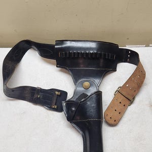 Op de afbeelding: Een zwart lederen pistoolholster bevestigd aan een brede riem met een reeks kleine lussen voor munitie. De holster is voorzien van een messingkleurige drukknoopsluiting en een secundaire riem. De riem heeft een beige verstelbaar gedeelte.