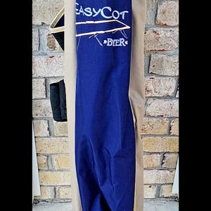 May include: A folded portable cot in a storage bag. The bag is primarily navy blue with tan side panels and features the white text "EASYCOT" above a graphic of a cot and the word "BYER". The base shows small grey metal feet at the bottom.