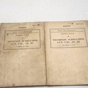 May include: Two off-white paper technical manuals titled War Department Technical Manual Thompson Submachine Gun, Cal. .45, M1, dated October 10, 1942. The covers are marked Restricted and TM 9-215, featuring black text on a weathered, aged surface.