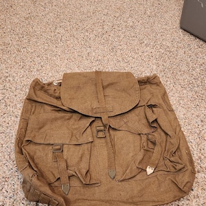 May include: A brown canvas rucksack featuring a large top flap with a central strap and buckle closure. The front includes two smaller pockets with flap covers, each secured by a strap and buckle. The bag has a textured fabric finish throughout.