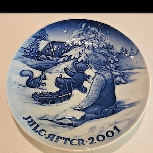 May include: A round ceramic decorative plate featuring a blue and white winter scene of a child playing in the snow with a teddy bear and a toy train. A squirrel is nearby, and a house sits in the background. The text JUL AFTEN 2001 is printed.