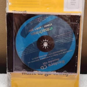 Könnte beinhalten: Eine versiegelte eMachines Restore-CD für den eTower 466ix 500i, bestehend aus einer blauen Disc mit weißem Text in einer transparenten Kunststoffhülle. Die CD ist mit einer gelben Karte verpackt, die oben rechts ein Feld für eine Briefmarke hat.
