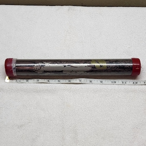 May include: A cylindrical storage tube with a metallic, weathered finish and dark red plastic end caps. A white measuring tape is positioned along the length of the tube, indicating a total length of approximately 12 inches from end to end.
