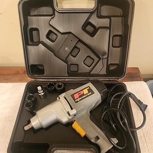 May include: A gray and black electric impact wrench with a yellow trigger, labeled PT Performance Tool, resting inside a black molded plastic carrying case. The set includes several black metal sockets arranged in the case alongside the power cord.