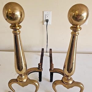 Puede incluir: Un par de morillos de chimenea de latón pulido con columnas altas y cónicas rematadas con grandes remates esféricos. Cada morillo se apoya sobre una base curva de tres patas con pies decorativos, conectada a una barra de soporte de hierro.