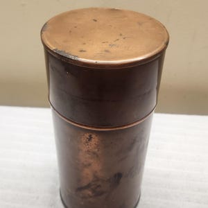 May include: A cylindrical copper canister with a matching lid. The metal surface features a warm, metallic brown tone with visible signs of natural oxidation, including dark spots and minor surface wear throughout the exterior body and lid.