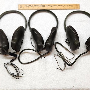 May include: Three sets of dark olive green over-ear headphones with adjustable headbands and attached black audio cables. A wooden ruler labelled North Carolina State Highway Patrol is positioned above the headphones for scale reference.