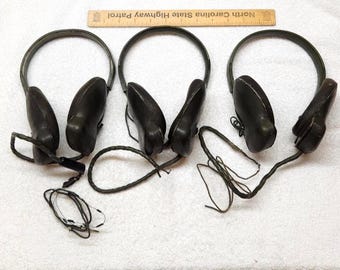 May include: Three sets of dark olive green over-ear headphones with adjustable headbands and attached black audio cables. A wooden ruler labeled North Carolina State Highway Patrol is positioned above the headphones for scale reference.