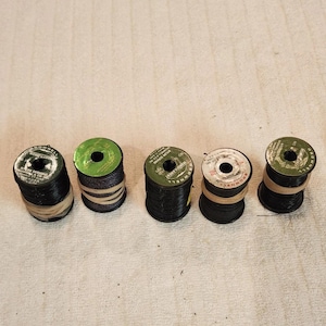 May include: Five cylindrical spools of black nylon serving thread arranged in a horizontal row. Each spool features a circular label on top with printed text including the brand name Brownell and product specifications, secured with rubber bands.