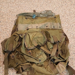 May include: An olive green canvas backpack featuring multiple exterior pockets with flap closures, adjustable webbing straps, metal grommets, and a drawstring top opening. The bag includes reinforced stitching and a weathered, textured fabric finish.