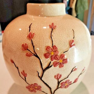 May include: A round ceramic vase with a cream-coloured crackle glaze finish. The vase features a hand-painted design of dark brown branches with small, vibrant pink blossoms and yellow centres, extending across the front of the rounded body.