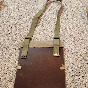 May include: A rectangular dark brown board with a smooth surface, framed by olive green canvas webbing. The item features adjustable olive green canvas straps attached with brass-colored metal hardware and two brass-colored metal studs at the base.