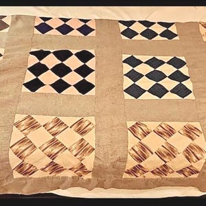 May include: A rectangular quilt featuring a grid of twelve square blocks separated by wide light gray fabric strips. Each square block contains a smaller diamond-patterned design in varying colors including brown, navy blue, and tan, with a textured finish.