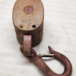 Puede incluir: Un bloque de polea de madera vintage con acabado desgastado, que presenta un emblema circular rojo central y un gancho de metal oxidado resistente unido a un soporte giratorio. El cuerpo de madera muestra signos de desgaste y orificios visibles.