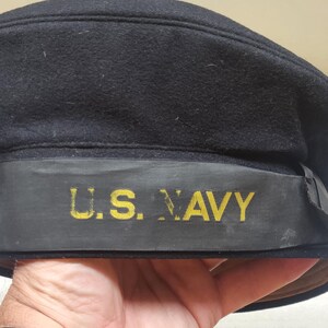 Pode incluir: Um gorro de marinheiro de lã azul-marinho escuro com uma faixa de fita preta em torno da base. A fita é bordada com o texto U.S. NAVY em letras maiúsculas amarelas em negrito. O gorro tem um topo plano e uma coroa estruturada.