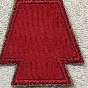 May include: A red embroidered patch in the shape of an arrowhead or a stylized shield. The patch features a solid red center with a thick, dark red stitched border and a light tan or beige merrowed edge finish around the entire perimeter.