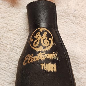 Könnte beinhalten: Eine schwarze, konisch zulaufende, flexible Gummihülle mit dem goldenen GE-Logo und dem aufgedruckten Text "Electronic TUBES" auf der Vorderseite. Das Objekt hat eine zylindrische Öffnung oben, die sich nach unten hin verbreitert.