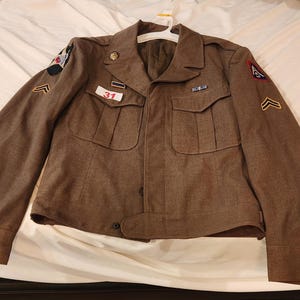 May include: A brown military-style jacket featuring a collared neckline, long sleeves with buttoned cuffs, and two front chest pockets. The jacket displays various patches, pins, and a white rectangular tag with the red number 31 printed on it.