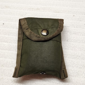Puede incluir: Una bolsa utilitaria rectangular de color verde oliva con una solapa plegable y un cierre de botón a presión de metal oscuro. La bolsa está fabricada con un tejido resistente con bordes reforzados de nailon y un pequeño broche a presión.