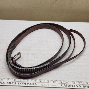 May include: A dark brown leather belt featuring a row of metallic cylindrical studs along the strap. The belt includes a rectangular metal buckle and is positioned next to a ruler showing a length of approximately 46 cm.