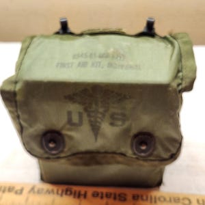 Puede incluir: Un estuche de primeros auxilios individual de color verde oliva con un símbolo de caduceo y las letras U S impresas en la solapa frontal. El texto 6545-01-068-8112 FIRST AID KIT, INDIVIDUAL es visible sobre el símbolo. Dos broches negros.
