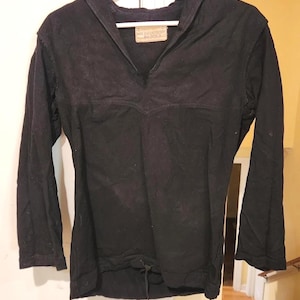 Puede incluir: Una camisa negra de manga larga con cuello estilo marinero, escote en V y dobladillo con cordón. La prenda incluye una etiqueta rectangular en el interior del cuello con texto visible y se muestra colgada en una percha de plástico blanca.