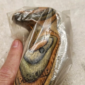 May include: A wooden pistol grip featuring a laminated wood grain pattern in shades of brown, tan, and black. The grip has a contoured shape with a single screw hole and the word PROLINE embossed in capital letters on the lower side surface.