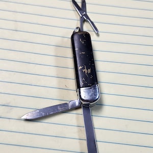 May include: A multi-tool pocket knife with a black handle showing signs of wear and gold-coloured flecks. The tool features a silver-toned metal casing, a small pair of extended scissors, a sharp blade, and a flat-head screwdriver attachment.