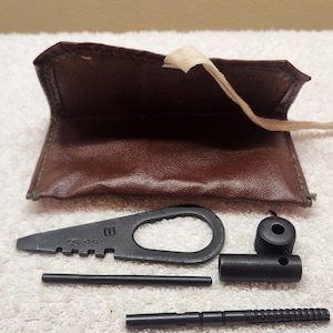 Könnte beinhalten: Ein brauner Lederbeutel mit Bindeband, begleitet von einem Set schwarzer Metallwerkzeuge, darunter ein flaches, spitzes Multifunktionswerkzeug mit der Aufschrift 75 95, ein dünner Stab, ein zylindrisches Teil mit Rändelknopf und ein gerillter Stab.
