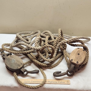 Puede incluir: Un sistema de poleas vintage que incluye dos bloques de madera con ganchos de metal oxidado y una cuerda gruesa de fibra natural enrollada. Una regla de madera con el texto North Carolina State Highway Patrol está colocada debajo.